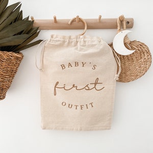 Puede incluir: Una bolsa de lona beige con cierre de cordón y una percha de madera. La bolsa tiene el texto "BABY'S first OUTFIT" impreso en marrón.