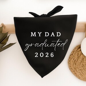 May include: Black triangular pet bandana with the text "MY DAD graduated 2026" in white script and block letters. The bandana is tied at the top with a black bow. The background is white.