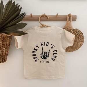 Pu&ograve; includere: Una maglietta color crema con una grafica nera di una mano scheletrica che fa il segno del rock and roll. La grafica &egrave; all'interno di un cerchio con il testo "Spooky Kid's Club Est 1642".