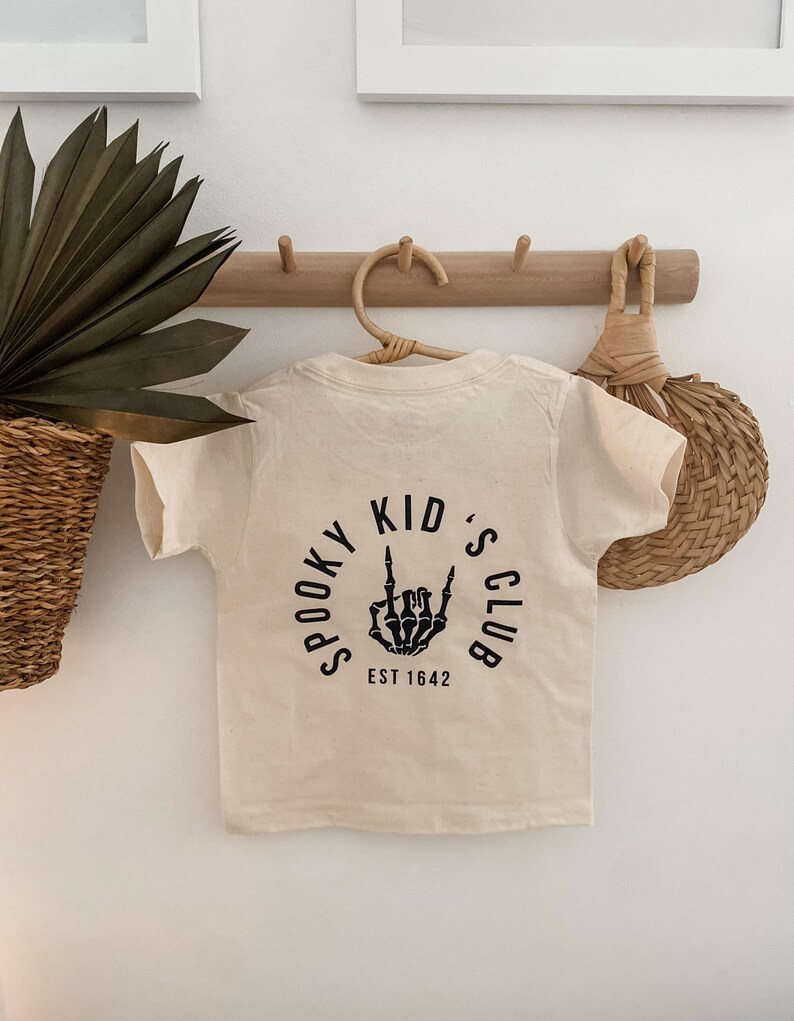Pu&ograve; includere: Una maglietta color crema con una grafica nera di una mano scheletrica che fa il segno del rock and roll. La grafica &egrave; all'interno di un cerchio con il testo "Spooky Kid's Club Est 1642".