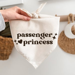 May include: Cream-colored dog bandana with the words "passenger princess" in a retro font, accompanied by star and heart graphics. The bandana is tied at the top and held by a hand.