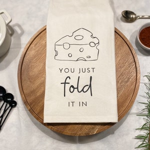 May include: A white kitchen towel with a black and white illustration of a wedge of cheese and the text "You just fold it in".