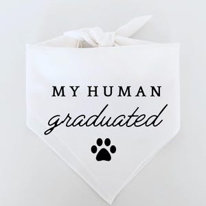 Puede incluir: Bandana blanca para perro con el texto "MY HUMAN graduated" en negro y una huella de pata negra. La bandana está anudada en la parte superior, formando un triángulo.