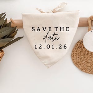 May include: Cream-colored triangular fabric with the text "SAVE THE date" and the date "12.01.26" in black. The fabric is hanging on a wooden rack, with a woven basket and a circular woven item in the background.