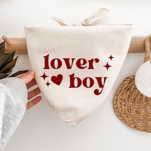 May include: A triangular off-white fabric pet bandana featuring the dark red text "lover boy" printed on the front. The design includes a small heart icon and four-pointed star accents. The bandana has fabric ties at the top for securing it.