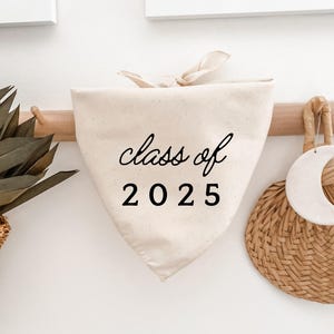 Puede incluir: Bandana crema para perro con el texto "class of 2025" en escritura negra. La bandana está atada en la parte superior y cuelga de una barra de madera. Una cesta tejida y elementos decorativos son visibles en el fondo.