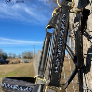 May include: A close-up shot of a black leather horse halter, featuring gold-coloured metalwork and a line of shimmering, iridescent crystals. The halter is shown outdoors against a blue sky and a wooden fence. The brand name "Crazy H Creations" is visible.