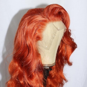 May include: A vibrant orange wig with long, wavy hair. The wig is styled with a side part and voluminous curls. It is displayed on a mannequin head against a white backdrop. The wig's color is a bright, fiery orange.