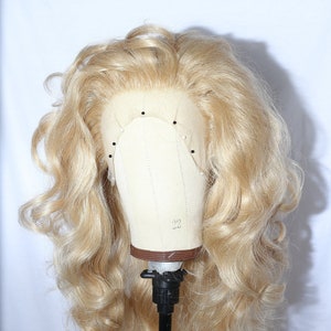 Puede incluir: Una peluca larga y rubia con rizos sueltos, peinada con un estilo ondulado y voluminoso. La peluca se muestra en una cabeza de maniquí con un soporte negro.