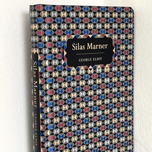 Op de afbeelding: Een hardcover boek getiteld "Silas Marner" van George Eliot. De boekomslag heeft een geometrisch patroon van blauwe, rode, gele en grijze ruiten. De titel en de naam van de auteur staan op een zwart label.