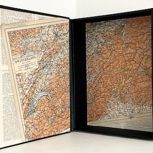 Può includere: Un libro svuotato con esterno nero e cornice interna nera. I pannelli interni presentano stampe di mappe vintage della Svizzera in tonalità marrone, beige e blu. Il lato sinistro mostra una mappa con il testo SWITZERLAND.