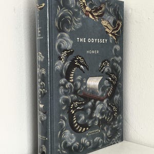 May include: A hardcover book titled The Odyssey by Homer from the Cranford Collection. The dark blue cover features gold and white illustrations of a ship surrounded by sea monsters and birds amidst swirling waves, with text on the spine.