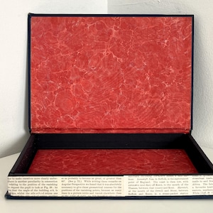 May include: Open rectangular box with a red marbled interior and a black exterior. The box's edges are trimmed with dark blue fabric. The base of the box is lined with text from a vintage book.