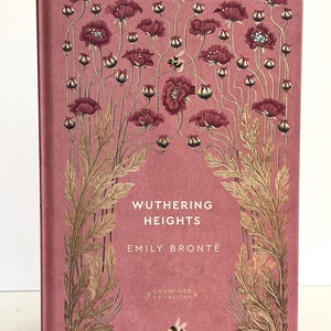 Pu&ograve; includere: Un libro con copertina rigida intitolato Wuthering Heights di Emily Bront&euml; della Cranford Collection. La copertina rosa polvere presenta illustrazioni dorate di alti papaveri, steli frondosi e diversi bombi sulla parte anteriore.