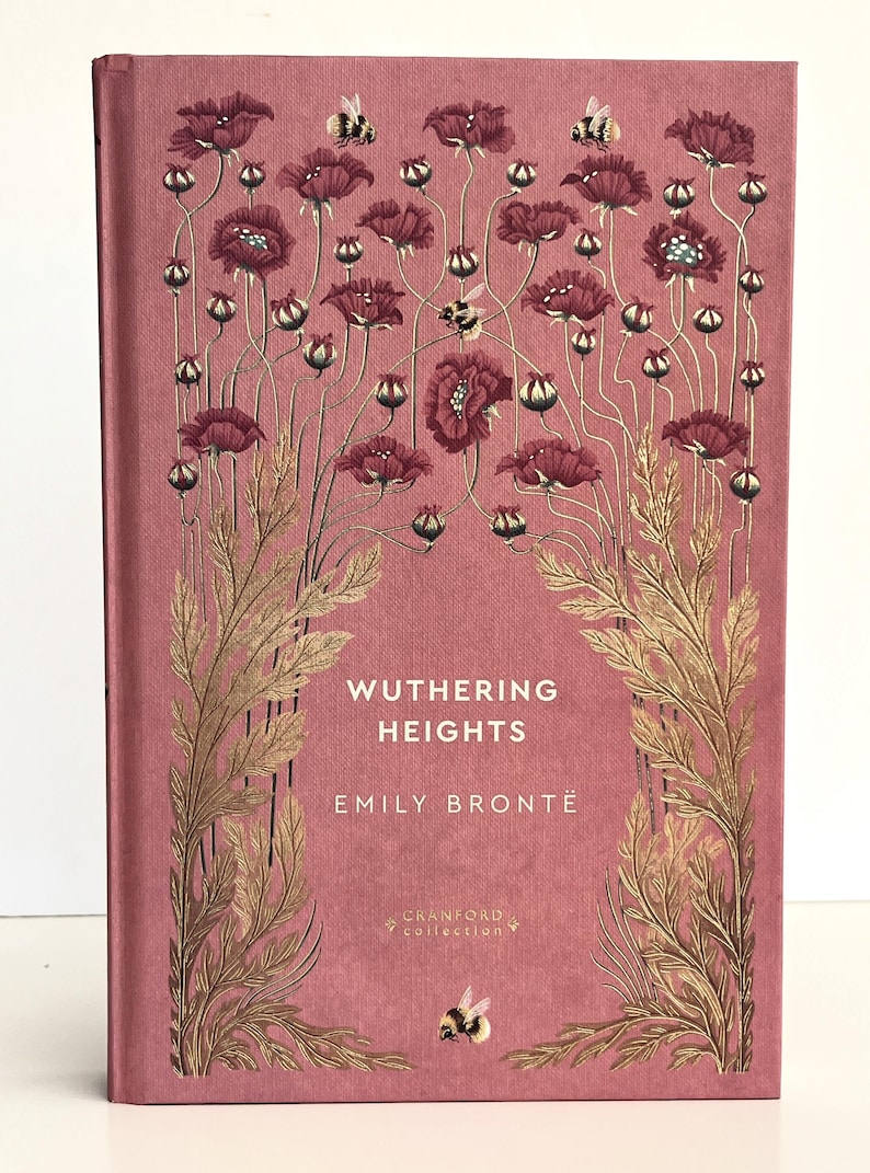 Pu&ograve; includere: Un libro con copertina rigida intitolato Wuthering Heights di Emily Bront&euml; della Cranford Collection. La copertina rosa polvere presenta illustrazioni dorate di alti papaveri, steli frondosi e diversi bombi sulla parte anteriore.