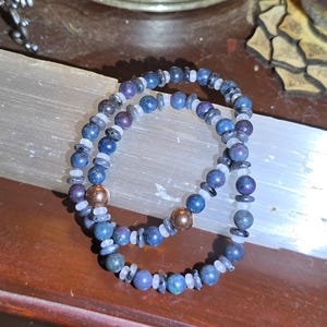 May include: A beaded bracelet featuring round blue and purple mottled stones, small translucent white spacer beads, thin dark grey disc-shaped beads, and two smooth, metallic copper-colored spherical beads arranged in a circular pattern.