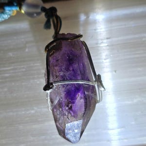 May include: A raw, deep purple amethyst crystal point pendant wrapped in dark copper and silver-toned wire. The crystal features natural facets and internal inclusions, suspended from a dark cord with a translucent, pale bead near the top.