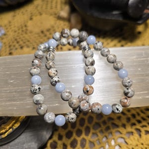 May include: Two beaded bracelets. The bracelets feature a mix of speckled, neutral-toned beads and light blue beads. The bracelets are displayed on a light-colored surface with a yellow crocheted background.