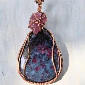 Peut inclure: Un pendentif en forme de goutte composé d'une pierre polie bleu foncé et magenta, entourée d'un fil de cuivre. Un petit cristal brut rose est fixé au sommet du pendentif, qui est suspendu à une chaîne en cuivre assortie.