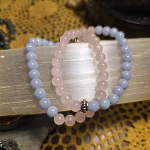 May include: Three beaded bracelets. One bracelet is light blue, another is pink, and the third is light blue. The bracelets are made of round beads and are displayed on a light-colored surface. The bracelets are a type of jewelry.