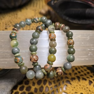 May include: Three beaded bracelets with a variety of green, brown, and tan beads. The bracelets are resting on a white rectangular object, with a yellow crocheted surface in the background. The beads have various patterns and sizes.