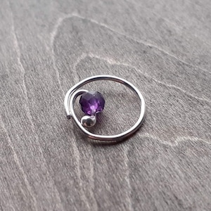 Earring with amethyst bead on a wooden background. The wire that forms this earring doubles over itself for about a sixth of the circumference. The other end of the wire is curled towards the center of the hoop and holds the amethyst bead.