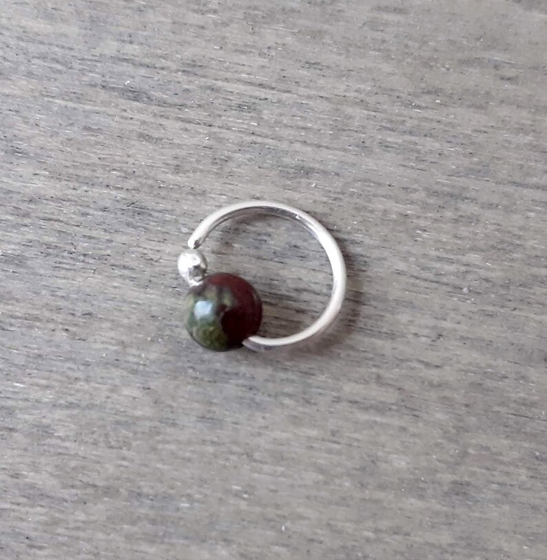 Dragons blood silver hoop earring against a wooden background. The hoop is made from a single piece of sterling silver balled at one end to hold the dragons blood bead in place. The bead is threaded through.