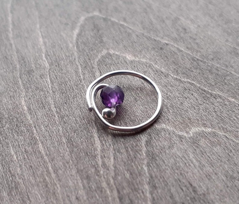 Earring with amethyst bead on a wooden background. The wire that forms this earring doubles over itself for about a sixth of the circumference. The other end of the wire is curled towards the center of the hoop and holds the amethyst bead.