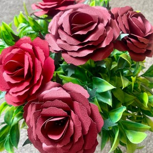 May include: A bouquet of five burgundy paper roses arranged in a green leafy bush. The roses are made of paper and have a textured, layered appearance.