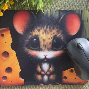 May include: Mouse mat featuring a cartoon mouse with big eyes, red ears, and a cheese design. A black wireless computer mouse is next to the mouse mat. The mouse mat has a black background.
