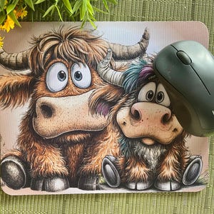 May include: A mouse mat with a cartoon illustration of two Highland cows with large eyes and shaggy brown fur. A dark grey wireless mouse rests on the mat. The mouse mat has a light brown background.