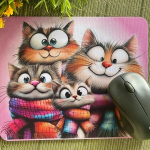 May include: A pink rectangular mouse mat with a cartoon illustration of four smiling cats. The cats have orange, brown, and grey fur and wear colourful scarves. A grey Logitech mouse rests on the mat.