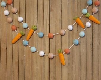 May include: A garland with felt balls in white, orange, and blue, with felt carrot decorations. The garland is hanging on a wood background.