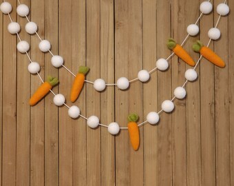 May include: Easter-themed garland featuring white felt balls and orange felt carrots with green tops. The garland is strung on white string and hangs against a wooden background, perfect for spring decor.