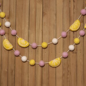 May include: A garland with pink, white, and yellow felt balls and lemon slices. The garland is strung on a white string and is hanging on a wooden background.