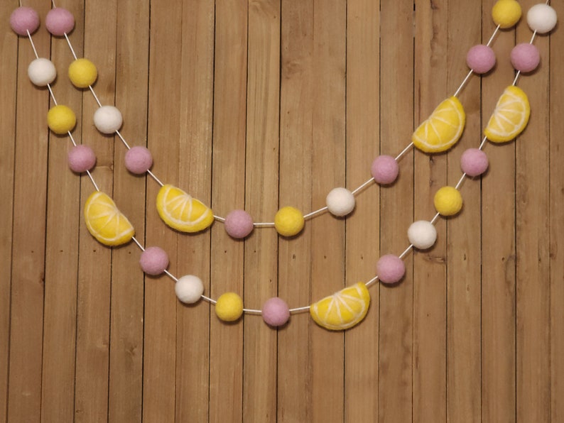 May include: A garland with pink, white, and yellow felt balls and lemon slices. The garland is strung on a white string and is hanging on a wooden background.