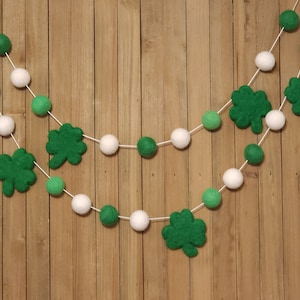 May include: Two strands of St. Patrick's Day garland. Each strand features white and green felt balls, and green felt shamrocks. The garland is strung on white string and hangs against a wooden background.