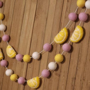 May include: A garland made of felt balls in white, yellow, and pink with lemon slices in between. The garland is strung on white twine and is hanging on a wood background.
