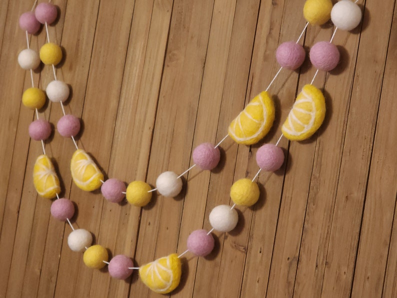 May include: A garland made of felt balls in white, yellow, and pink with lemon slices in between. The garland is strung on white twine and is hanging on a wood background.