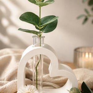 May include: A white heart-shaped frame holding a clear glass test tube vase. A single green eucalyptus stem with small leaves and a light-coloured flower bud is placed inside the tube. Two small white daisy-like flowers rest at the base of the frame.
