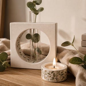 May include: A square off-white ceramic vase with a circular cutout containing a clear glass test tube holding a green eucalyptus stem. Beside it sits a small round ceramic candle holder with a floral pattern and a lit white tea light candle.