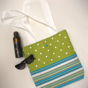 Puede incluir: Bolso tote de lunares verdes y blancos con un fondo a rayas azules y blancas. El bolso tiene asas blancas y está sobre una superficie blanca. Hay gafas de sol y una botella de protector solar al lado del bolso.