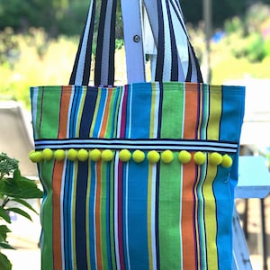 Puede incluir: Una bolsa de mano a rayas con fondo azul brillante y ribete de pompones amarillos. La bolsa tiene asas rayadas en blanco y negro.