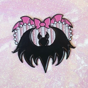 Puede incluir: Una silueta de murciélago negro con alas de purpurina rosa y blanca y un lazo rosa en la parte superior. El murciélago tiene forma de corazón.