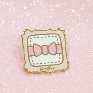 Puede incluir: Un pin de esmalte de color dorado con un cuadrado blanco que contiene un lazo rosa en su interior. El cuadrado tiene un borde blanco con una línea punteada y el texto "Dollface" está escrito debajo del cuadrado.
