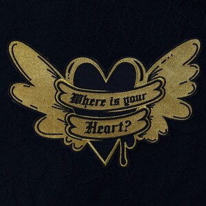 Puede incluir: Parche bordado en oro con un corazón negro y alas. El parche tiene el texto "Where is your Heart?" en una pancarta.