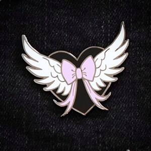 Puede incluir: Corazón negro con alas blancas y un lazo rosa. El corazón está bordeado de plata y las alas son blancas con un contorno plateado. El lazo es rosa con un contorno plateado.