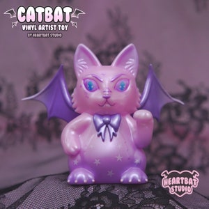 May include: A purple and pink vinyl toy cat with bat wings. The cat has a bow tie and stars on its body. The toy is called "Catbat" and is a vinyl artist toy by Heartbat Studio.