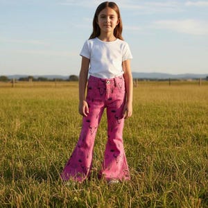Könnte beinhalten: Eine Person trägt ein weißes T-Shirt und eine rosa Schlaghose. Die Jeans haben ein schwarzes Muster aus Federn und Bullenschädeln. Die Person steht auf einer Wiese unter blauem Himmel.