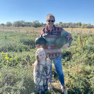 May include: A woman and a young girl are standing in a field, holding pumpkins. The woman is wearing a red and black patterned shirt, blue jeans, and sunglasses. The girl is wearing a brown faux fur vest over a floral dress.